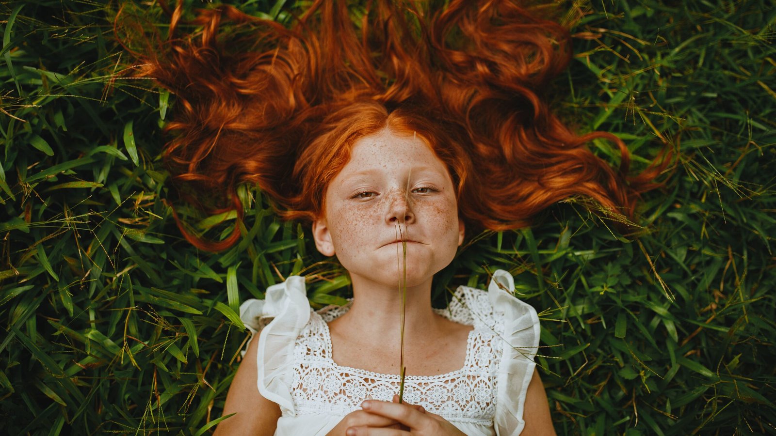 A red-haired girl in a white lace dress lying on green grass and holding a plant stem