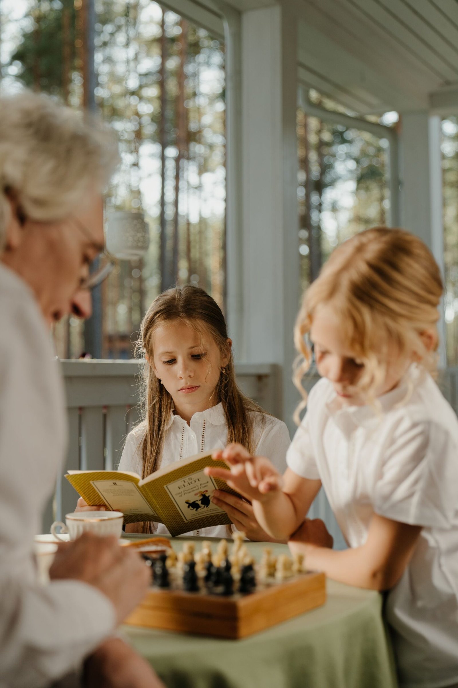 A young girl reading a book while sitting at a table with an adult and another child playing a chess game