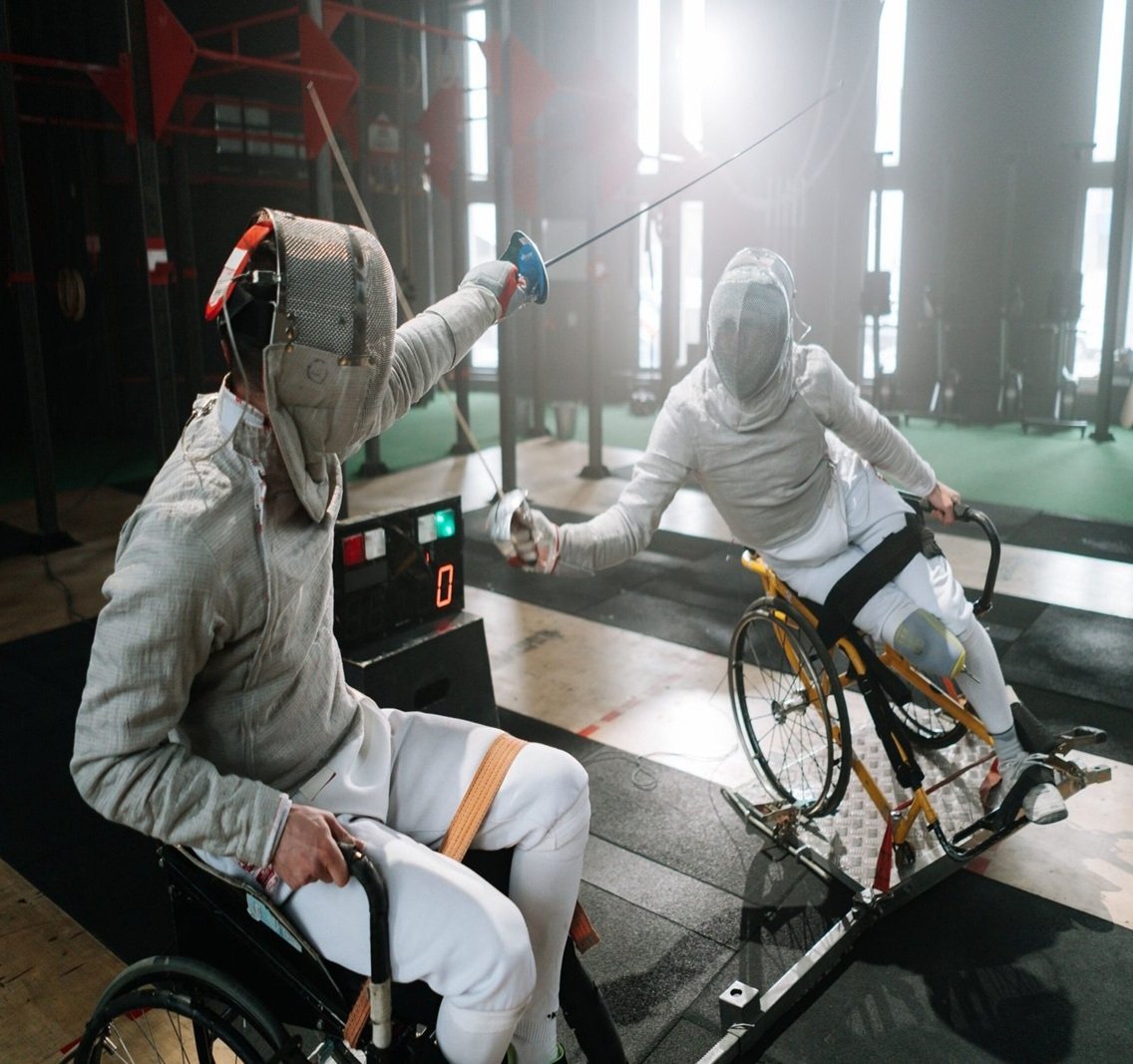 Two athletes competing in wheelchair fencing while wearing protective fencing masks and jackets